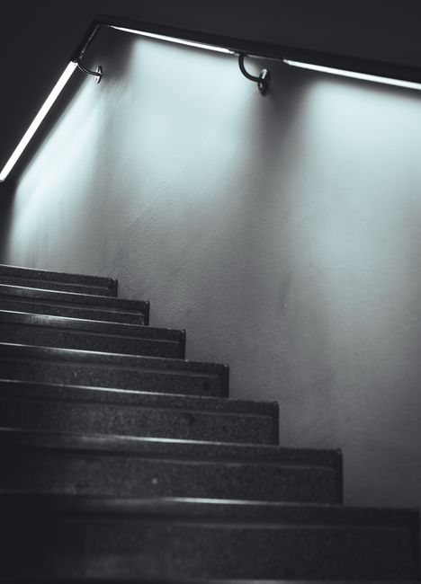 A downward view of a staircase inside a residential property with green carpeted steps and a dark wooden banister supported by turned wooden spindles. The staircase curves at the bottom, leading to an
