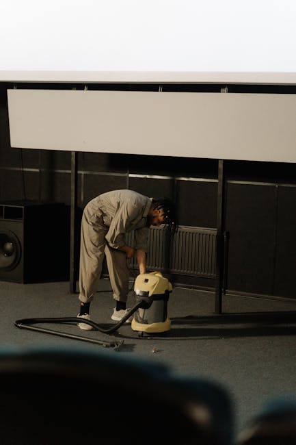 A person dressed in a light-colored jumpsuit is operating a yellow professional carpet cleaner vacuum on a carpeted floor in an indoor setting, likely a conference or event space, with dark wall panels and a large white projector screen or banner overhead. The individual is bent over, focusing on deep cleaning the carpet surface, which appears clean but may be in the process of being thoroughly sanitised or maintained. Visible equipment includes the vacuum hose and a speaker placed against the wall. Natural lighting illuminates the scene, highlighting the meticulous cleaning activity carried out by Carpet Cleaning Hounslow in line with end of tenancy cleaning standards, ensuring hygienic and well-maintained surfaces, suitable for both residential and commercial environments.