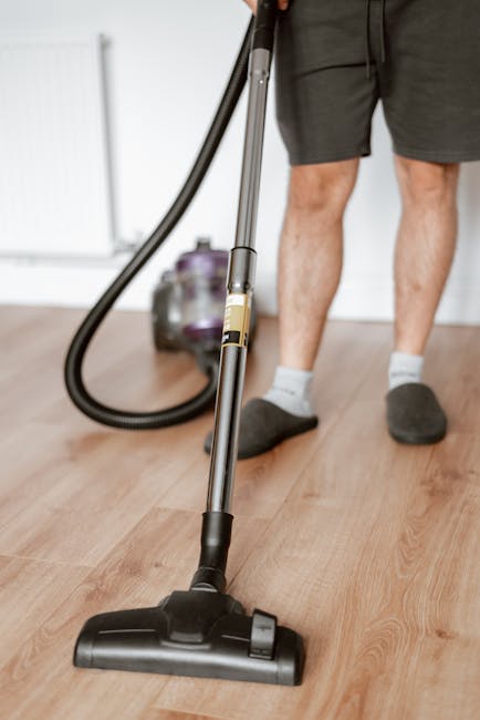 A person using a vacuum cleaner with a black hose and metallic silver wand to perform surface cleaning on a wooden floor in a residential room. The individual is wearing grey shorts, dark slippers, and white socks. The wooden floor appears clean and polished, reflecting some natural light. In the background, a purple and grey vacuum attachment is visible, resting on the floor. The scene showcases a professional approach to deep cleaning in a home setting, emphasizing hygiene and maintenance, consistent with services offered by Carpet Cleaning Hounslow as highlighted on their website for end of tenancy cleaning in Lampton Road, Hounslow.