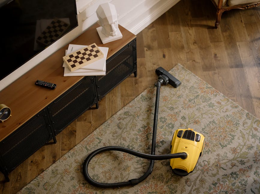 A person cleaning a carpet with a yellow and black vacuum cleaner on a patterned area rug in a well-lit room. The individual, wearing casual clothing including blue jeans and a beige top, is using a w