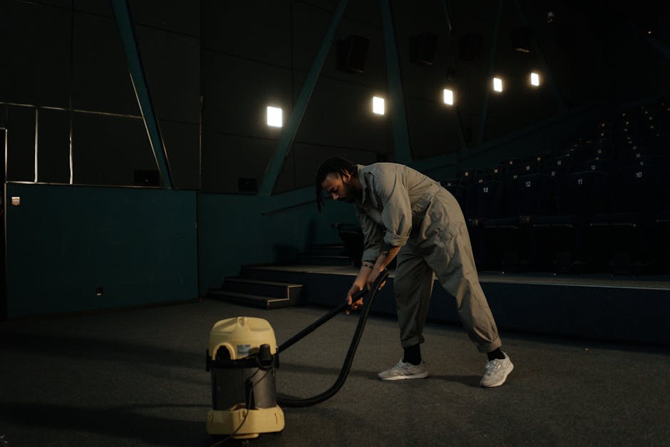 A woman using a yellow vacuum cleaner to deep clean a patterned beige and green carpet in a living room area, with visible wooden flooring surrounding the carpet and a red leather sofa nearby. The roo