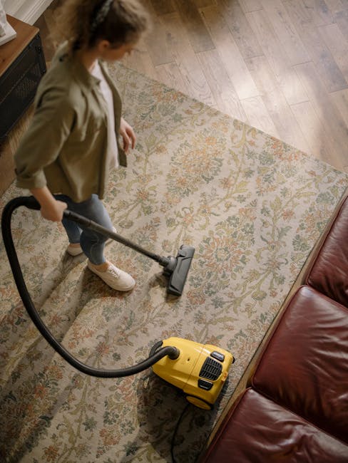 A person using a yellow vacuum cleaner to deep clean a patterned beige and pastel rug on a wooden floor in a living room, with a brown leather sofa nearby. The room is well-lit with natural light, and the surfaces appear clean and dust-free. The scene illustrates professional surface cleaning carried out by Carpet Cleaning Hounslow, highlighting thorough domestic and commercial upholstery maintenance.